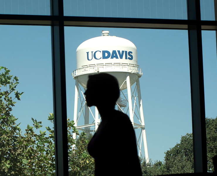 Silhouette of a person walking past a window, with the UC Davis water tower against a blue sky outside.