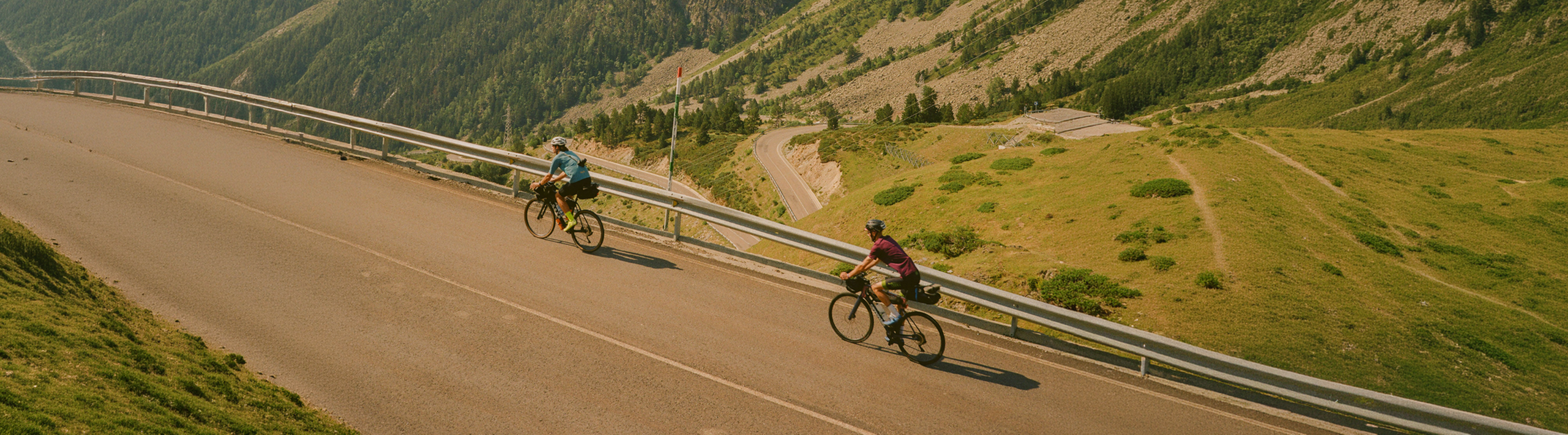 Two cyclists ride along a winding mountain road with grassy hills and trees in the background.