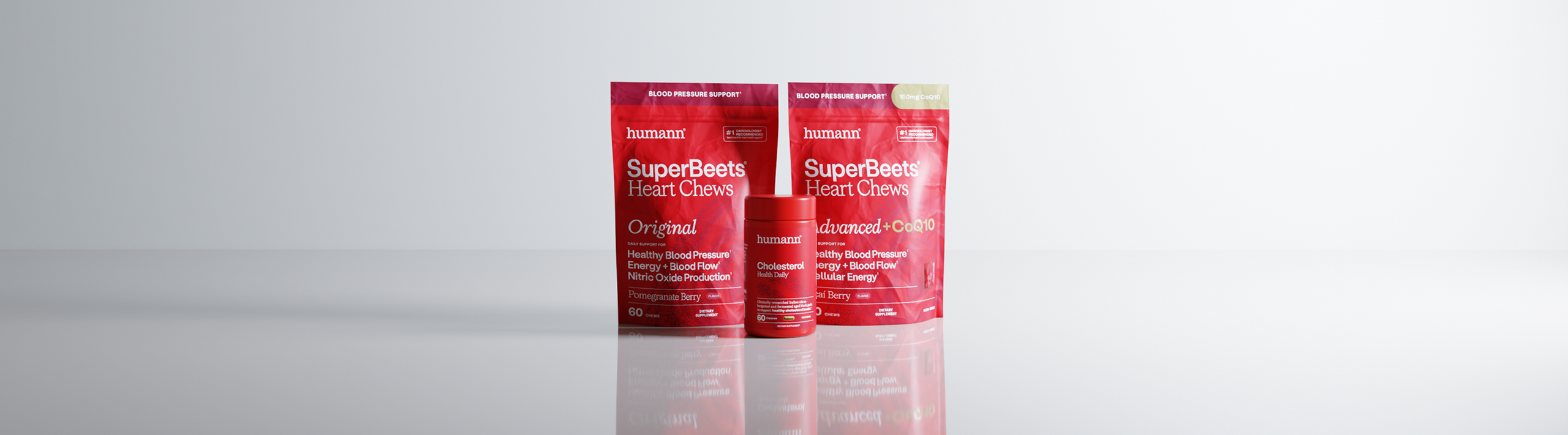 Three red Humann SuperBeets Heart Chews supplement packages on a light gray reflective background: one large pouch labeled Original, one large pouch labeled advanced+CoQ10, and one smaller cylindrical jar labeled Cholesterol Health Daily.