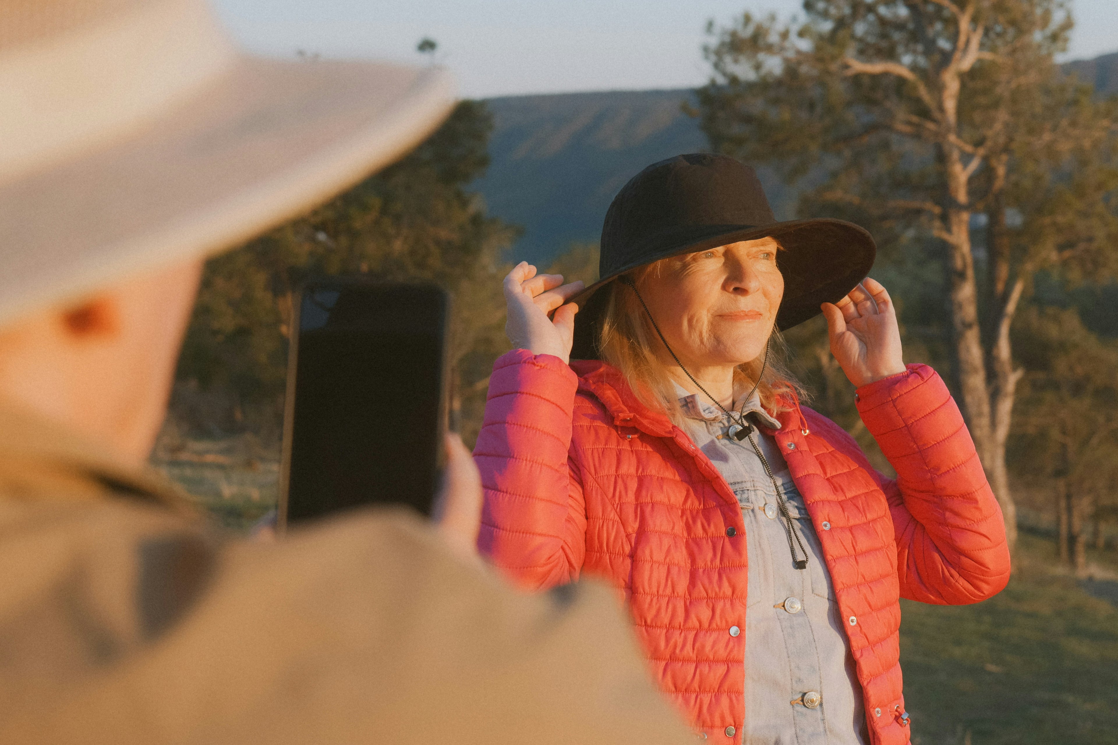 A woman outdoors wearing a black wide-brim hat and a bright pink puffer jacket, adjusting the brim of the hat while another person in the foreground takes a photo with a smartphone.