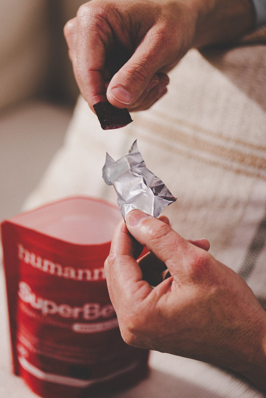 A close-up lifestyle shot of a person's hands unwrapping a silver foil packet and placing a dark red chewable tablet into it, with a red pouch of Humann SuperBeast energy powder in the foreground.