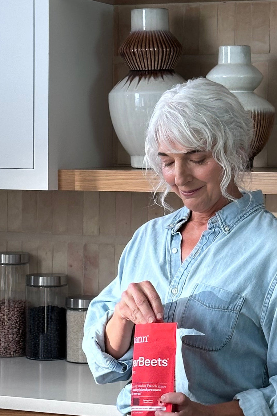 A woman with white hair stands in a modern kitchen, holding a red box of coffee. The kitchen features light wood cabinets, open wooden shelves with decorative items, and a marble countertop.
