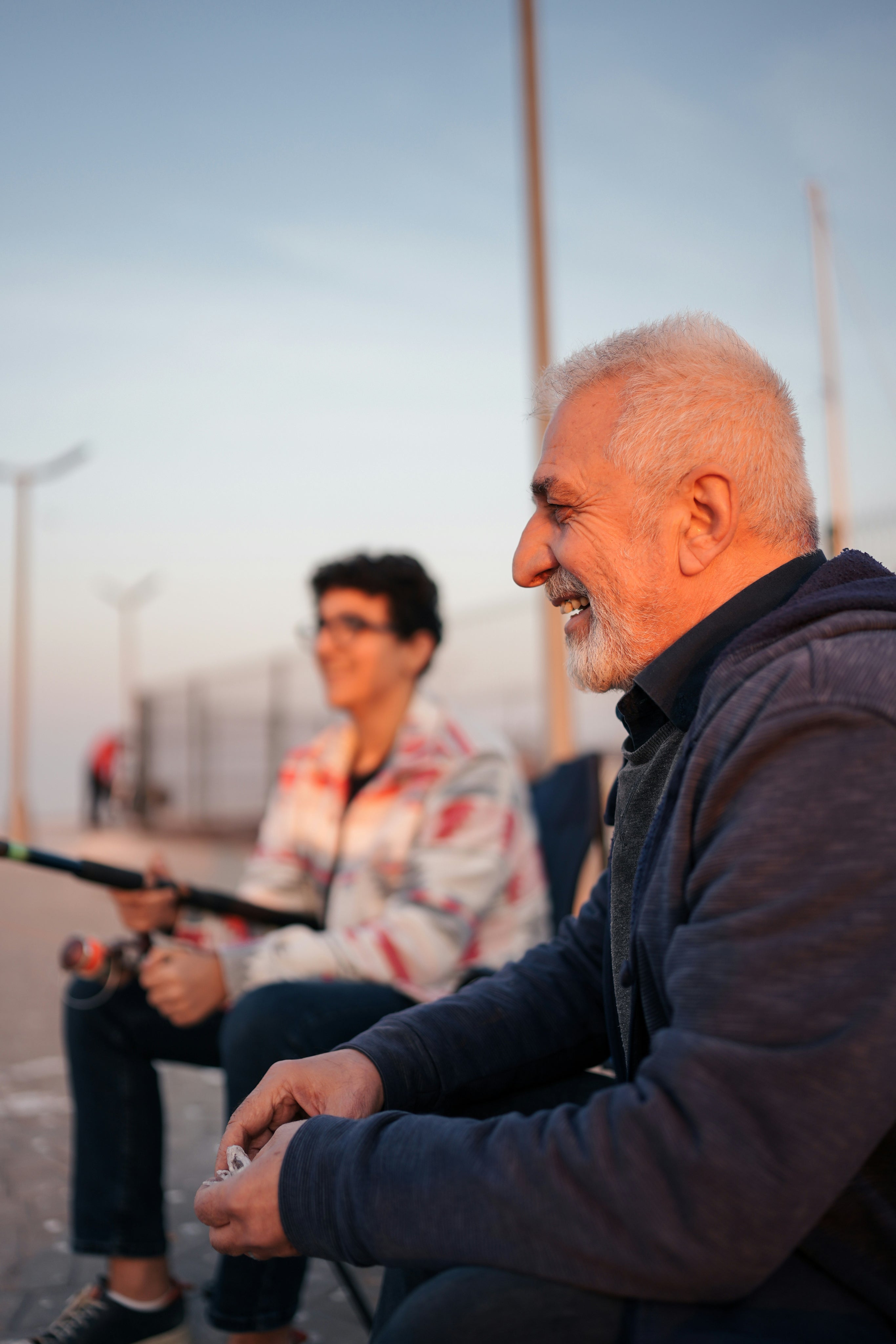 An older man with white hair and a beard sits in the foreground outdoors, smiling and looking to the side, while a woman in a patterned jacket sits blurred in the background holding a fishing rod.