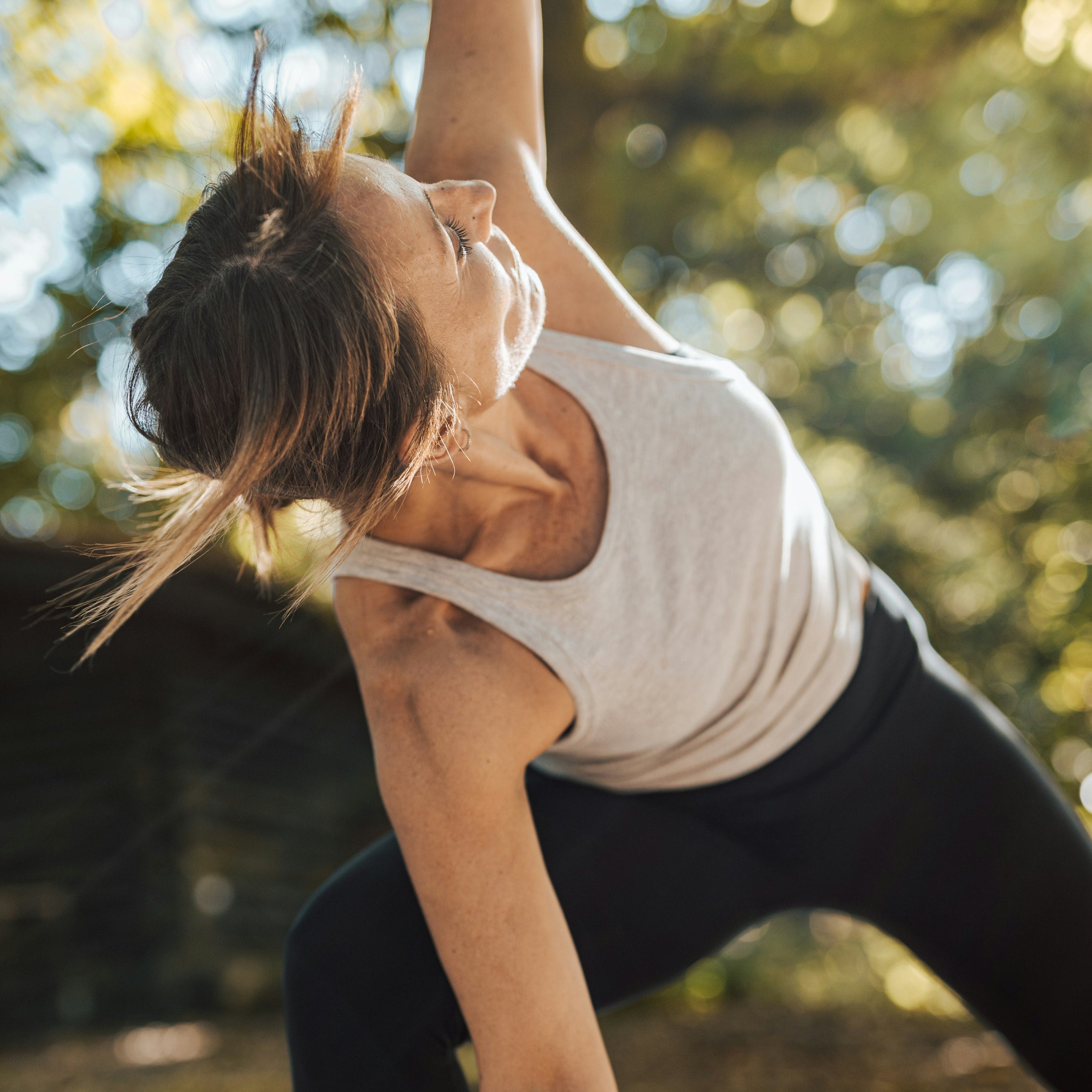 A woman in a white tank top and black leggings performs a yoga pose outdoors