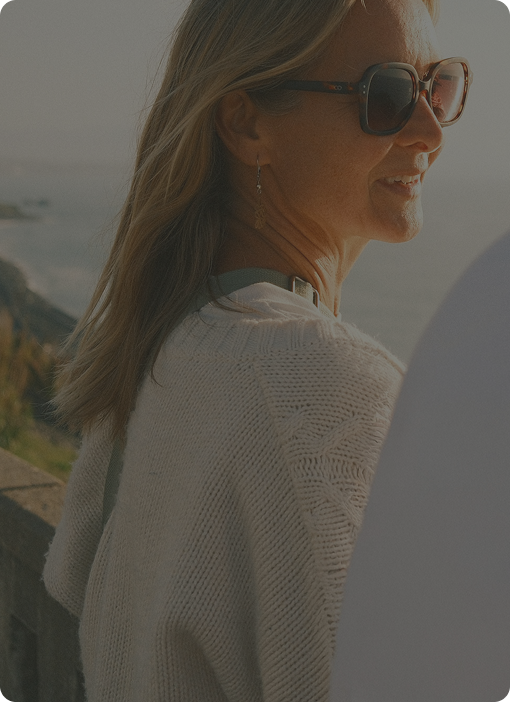 Woman in sunglasses and a cream sweater smiling by the coast